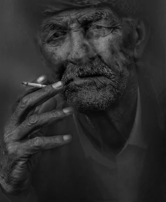 Portrait en noir et blanc d'un homme âgé au visage marqué portant un chapeau, tenant une cigarette allumée à la bouche avec de la fumée qui s'échappe autour de ses traits