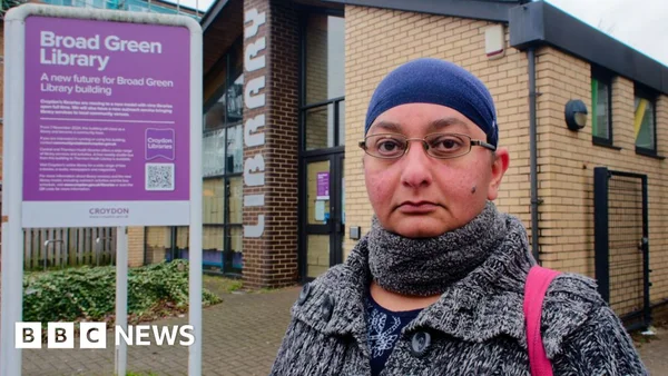 Une femme au foulard bleu et vêtement gris se tient devant la bibliothèque Broad Green, avec un panneau annonçant son avenir à sa gauche