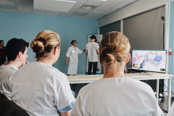Étudiants en soins infirmiers en uniformes blancs assis en salle de classe, observant une présentation sur un écran lors d'un cours de simulation numérique