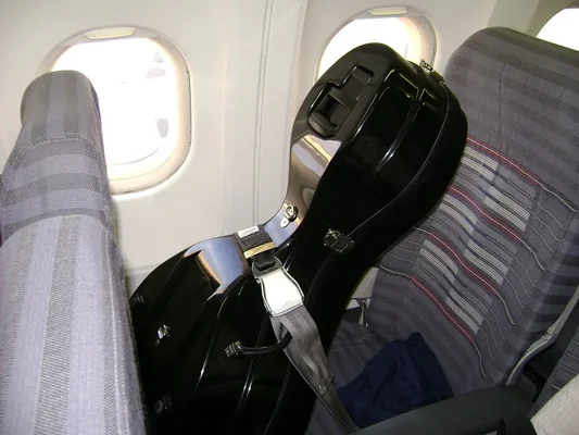 Un grand étui noir rigide pour instrument à cordes attaché par sa ceinture de sécurité sur un siège d'avion, près d'un hublot de la cabine aérienne