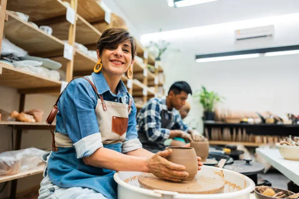 Une femme souriante en chemise bleue et tablier façonne une pièce d'argile sur un tour de potier dans un atelier de céramique, tandis qu'un autre artisan travaille en arrière-plan