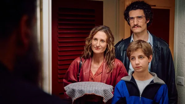 Camille Cottin, Louis Garrel et Simon Boublil dans une scène du film Juste une illusion : la femme tient un plat couvert devant une porte, l'homme à lunettes et moustache derrière elle, le jeune garçon en veste bleue devant