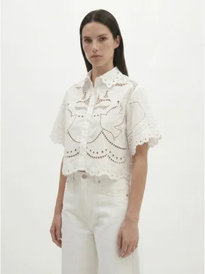 Femme portant un chemisier blanc à manches courtes avec broderies ajourées et jean blanc sur fond blanc uni