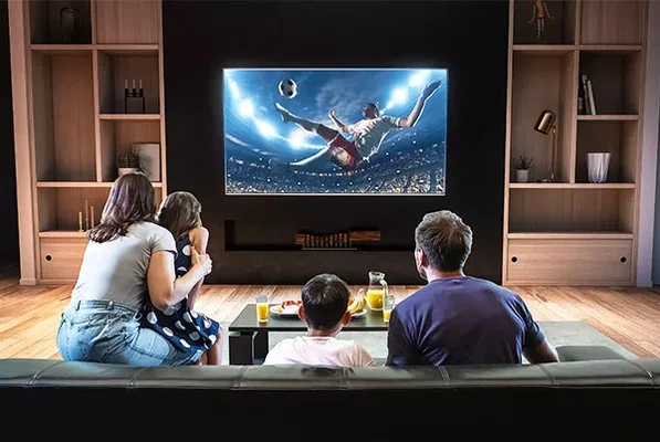 Une famille de quatre personnes assise sur un canapé dans un salon regarde un match de football sur un grand téléviseur mural à écran plat