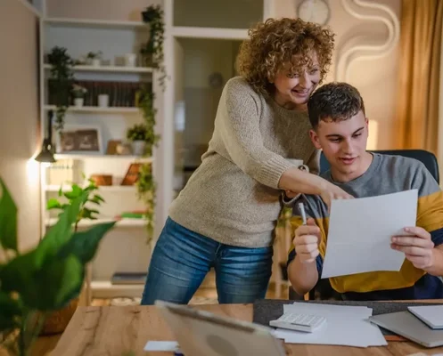 Une femme aux cheveux bouclés montre du doigt un document tenu par un jeune homme assis à son bureau, entouré d'un ordinateur portable et d'une calculatrice, illustrant les démarches bancaires pour un prêt étudiant