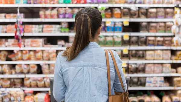 Une femme de dos, cheveux attachés en queue de cheval et vêtements décontractés, se tient avec un chariot devant les rayons d'alimentation d'un supermarché