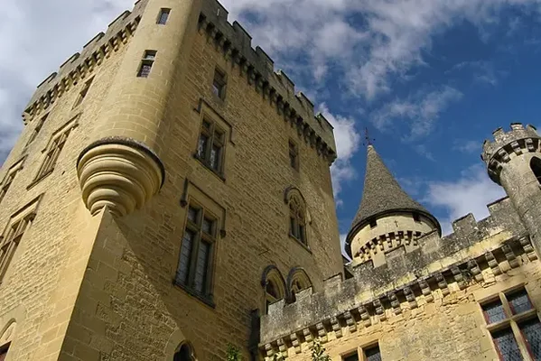Le château de Puymartin en Dordogne, forteresse médiévale en pierre avec une grande tour carrée à mâchicoulis et une tour conique sous un ciel bleu nuageux
