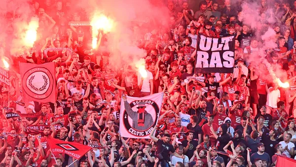 Supporters du Stade Rennais en tribune au Roazhon Park, illuminés par des fumigènes rouges, brandissant des drapeaux et bannières dont une portant l'inscription 'ULT RAS'