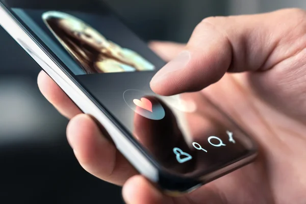 Doigt pressant l'icône en forme de cœur lumineuse sur l'écran d'une application de rencontres tenue dans la main, illustrant l'usage d'apps de rendez-vous au Japon