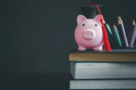 Tirelire rose coiffée d'un bonnet de diplômé posée sur une pile de livres, avec des crayons de couleur en arrière-plan, symbolisant l'épargne pour financer ses études supérieures