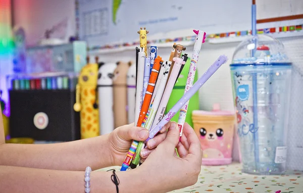 Des mains tenant une collection de stylos Legami colorés à personnages au-dessus d'un bureau avec de la papeterie en arrière-plan