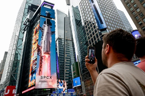 Un homme en t-shirt beige photographie au smartphone une grande affiche numérique SpaceX affichant un lancement de fusée et la mention « SPCX NasdaqListed », le jour de l'introduction en bourse de SpaceX au Nasdaq MarketSite de New York.