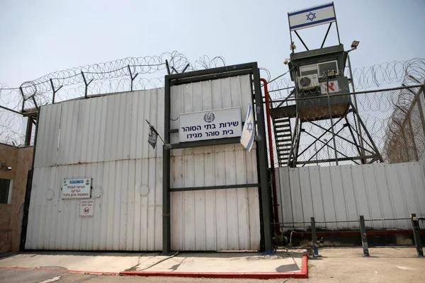 Entrée sécurisée d'une prison israélienne avec portail métallique portant une inscription en hébreu, fil barbelé et tour de guet arborant le drapeau israélien