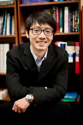 Jianyu Li, chercheur à l'Université McGill, portant des lunettes et un blazer sombre, les bras croisés, souriant devant une étagère de bois remplie de livres
