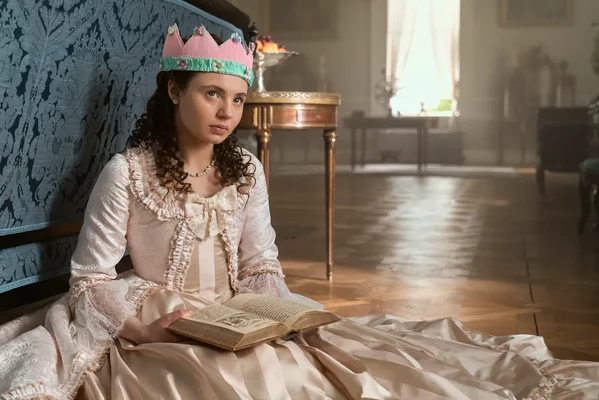 Jeune Violet de Bridgerton enfant, cheveux bouclés et couronne de papier rose, assise au sol d'un grand salon avec un livre ouvert sur les genoux