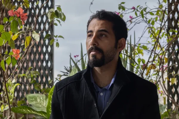 Le poète gazaoui Mosab Abu Toha, portant une veste noire sur une chemise bleue, debout en extérieur devant une clôture ajourée entourée de verdure et de fleurs
