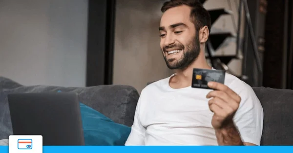 Homme barbu portant un t-shirt blanc, assis sur un canapé souriant, tenant une carte bancaire à la main devant un ordinateur portable ouvert