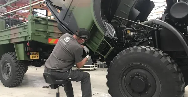 Un mécanicien en t-shirt gris et casquette assis sur un tabouret roulant travaille sur le moteur d'un camion militaire dont la cabine est basculée en avant dans l'atelier de l'usine Arquus de Saint-Nazaire.