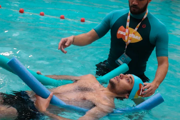 Une monitrice de natation en chemise de bain turquoise assiste un adulte flottant sur le dos dans une piscine à l'aide de flotteurs en mousse bleus