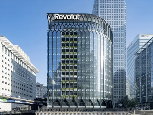 Immeuble de bureaux moderne au verre incurvé portant le logo Revolut en haut, situé à Canary Wharf parmi d'autres gratte-ciels et un pont ferroviaire à Londres