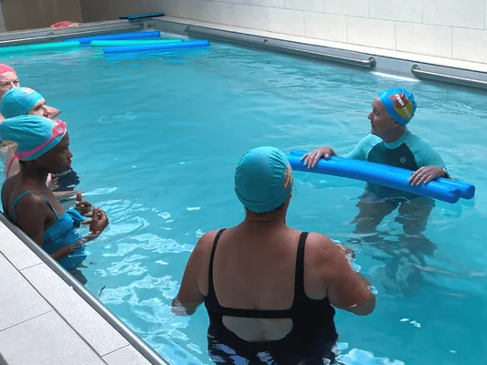 Un groupe de personnes portant des bonnets de natation dans une piscine intérieure, dont l'une tient un flotteur bleu et semble encadrer les autres en cours de natation