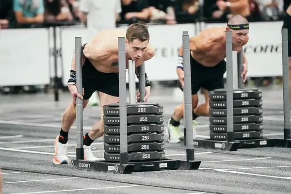 Deux athlètes masculins torse nu poussent des traîneaux chargés de disques de fonte sur la piste d'une épreuve Hyrox