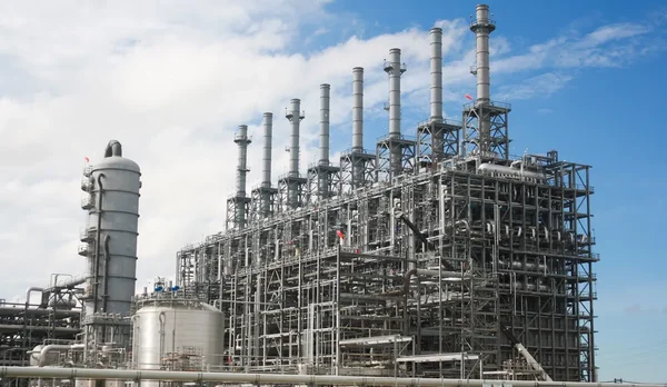 Vaste usine chimique avec ses structures d'acier, réseaux de tuyauteries et plusieurs cheminées industrielles s'élevant vers un ciel bleu nuageux