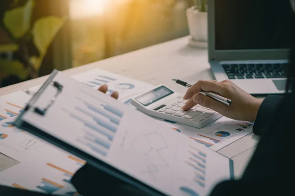 Mains d'une personne utilisant une calculatrice et un stylo sur des graphiques et tableaux financiers, avec un ordinateur portable en arrière-plan