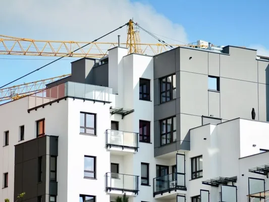 Façade d'un immeuble d'habitation moderne blanc et gris avec balcons, sous un ciel bleu, avec une grue de chantier jaune visible en arrière-plan