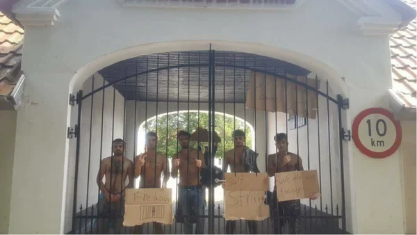 Cinq hommes sans chemise derrière une grille métallique noire d'un centre de détention pour expulsion au Danemark, tenant des pancartes en carton portant les mots « Freedom » et « Strike »