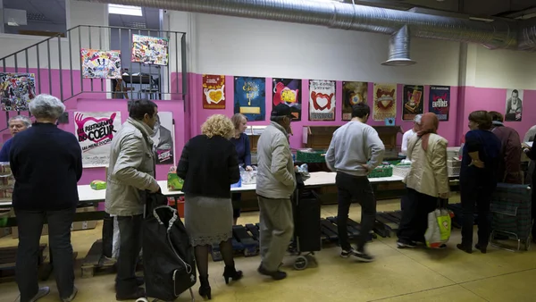 File de personnes devant de longues tables dans une grande salle aux murs roses avec affiches, accueillies par les Restos du Cœur lors d'une distribution alimentaire
