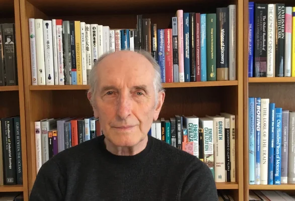 Portrait de l'analyste et essayiste tchèque-canadien Vaclav Smil, âgé, cheveux gris, portant un pull noir, assis devant une bibliothèque en bois remplie de livres