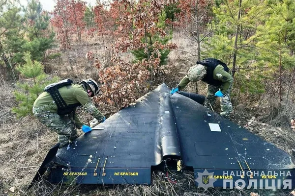 Deux individus en tenue militaire et gants bleus examinant l'épave d'un drone à voile delta noir, dénommé Geran-2 (version russe du Shahed-136), dans une zone boisée en Ukraine, avec un filigrane de la police nationale ukrainienne