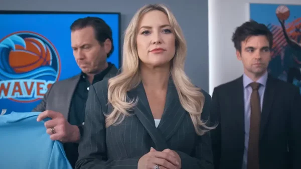 Kate Hudson en costume rayé dans Running Point, flanquée de deux collègues masculins devant un logo de franchise sportive sur Netflix