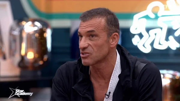 Christophe Pinna, ancien professeur de la Star Academy, portant une veste noire sur chemise blanche, lors d'une interview au logo Star Academy visible