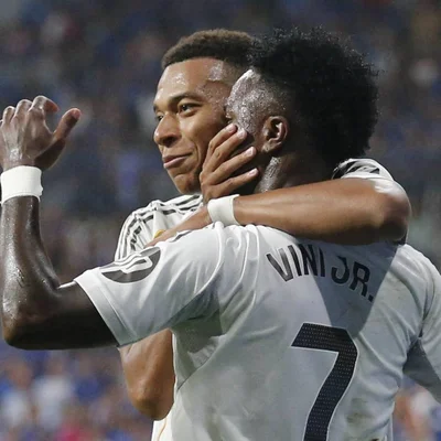 Kylian Mbappé et Vinicius Junior, en maillot blanc du Real Madrid, enlacés sur la pelouse, l'un posant son bras autour du cou de l'autre et sa main sur sa joue