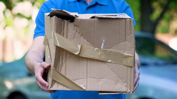 Personne portant une chemise bleue tenant un colis en carton endommagé scotché, illustrant une réception de colis abîmé