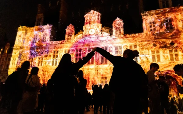 Foule de spectateurs en silhouette regardant une projection lumineuse colorée sur la façade d'un grand bâtiment parisien lors de la Nuit Blanche, deux figures centrales levant les bras en l'air