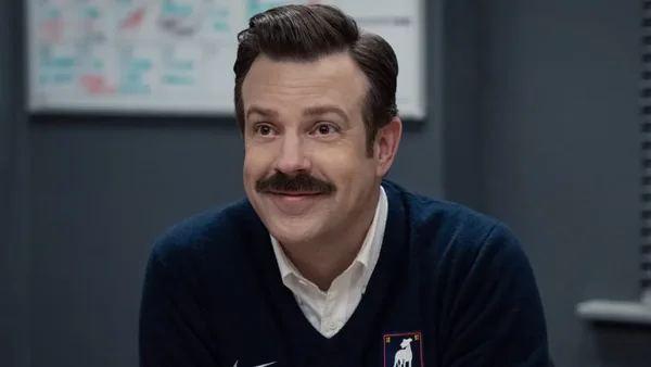 Jason Sudeikis, créateur et acteur principal de Ted Lasso, portant un pull marine sur chemise blanche, souriant légèrement en regardant hors champ
