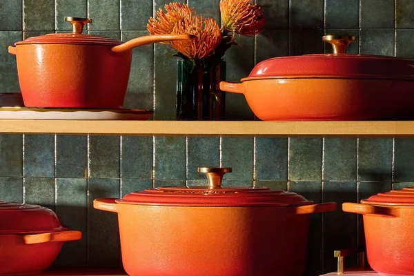 Collection de cocottes en fonte émaillée orange Le Creuset disposées sur deux étagères en bois devant un carrelage vert foncé, avec un vase de fleurs orange sur l'étagère supérieure