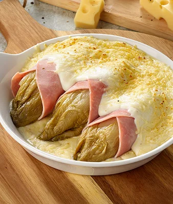 Plat blanc en céramique contenant trois endives au jambon recouvertes de sauce au fromage fondue, posé sur une planche en bois avec des morceaux de fromage en arrière-plan