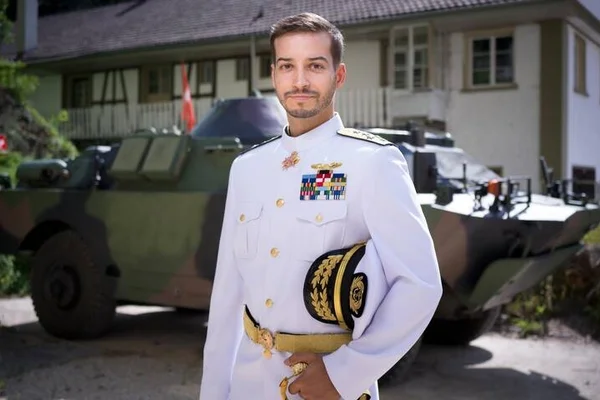 Jonas Lauwiner en uniforme militaire blanc brodé d'or et décoré de médailles, debout devant un véhicule blindé camouflé et un bâtiment