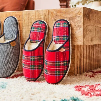 Paire de charentaises retro en laine kaki et une autre en carreaux rouge et gris, appuyées contre une table en bois sur un tapis à motif