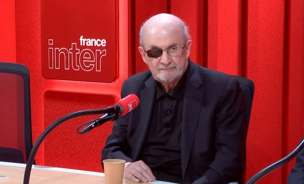 L'écrivain Salman Rushdie, barbe blanche et lunettes, en interview dans le studio de France Inter devant un fond rouge orné du logo de la radio