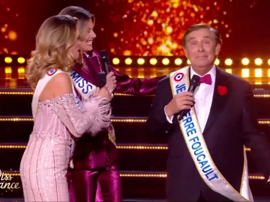 Jean-Pierre Foucault en smoking et ceinture portant le nom "Jean-Pierre Foucault", présentant Miss France 2026 sur scène aux côtés de deux femmes en tenues élégantes, devant un décor scintillant