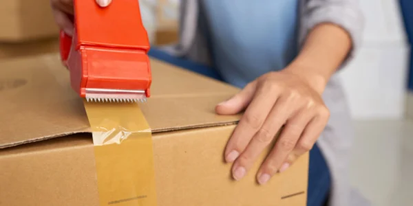 Gros plan sur les mains d'une personne utilisant un distributeur de ruban adhésif rouge pour sceller un carton de déménagement avec du ruban de packaging transparent