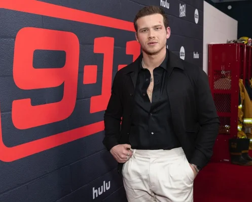 L'acteur britannique Oliver Stark, qui joue Buck dans la série 9-1-1, posant devant un panneau portant le logo de la série et la mention Hulu, vêtu d'une veste noire sur un t-shirt noir et un pantalon blanc