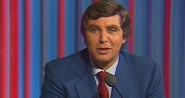 Jean-Pierre Foucault portant un costume bleu et une cravate rouge, parlant dans un micro devant un fond à rayures rouge et bleu, lors d'une rétrospective de sa carrière télévisuelle
