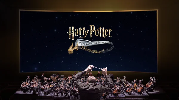 Un chef d'orchestre dirige un orchestre symphonique lors du Harry Potter Film Concert Series devant un grand écran affichant le logo de l'événement sur fond étoilé