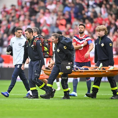 Tiago Santos blessé, évacué sur civière par le personnel médical du LOSC lors d'un match de Ligue 1, tandis qu'un coéquipier en maillot rouge observe depuis la pelouse